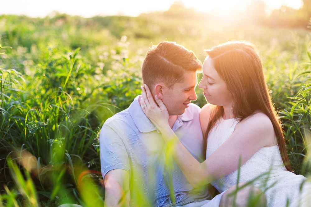 A couple shares an intimate moment sitting together in a sunlit field surrounded by green vegetation at sunset.