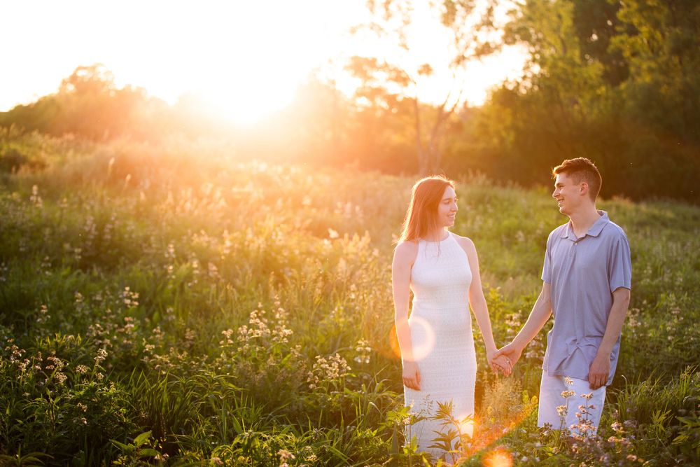 A romantic couple holds hands in a sunlit meadow during golden hour among wildflowers and tall grass.