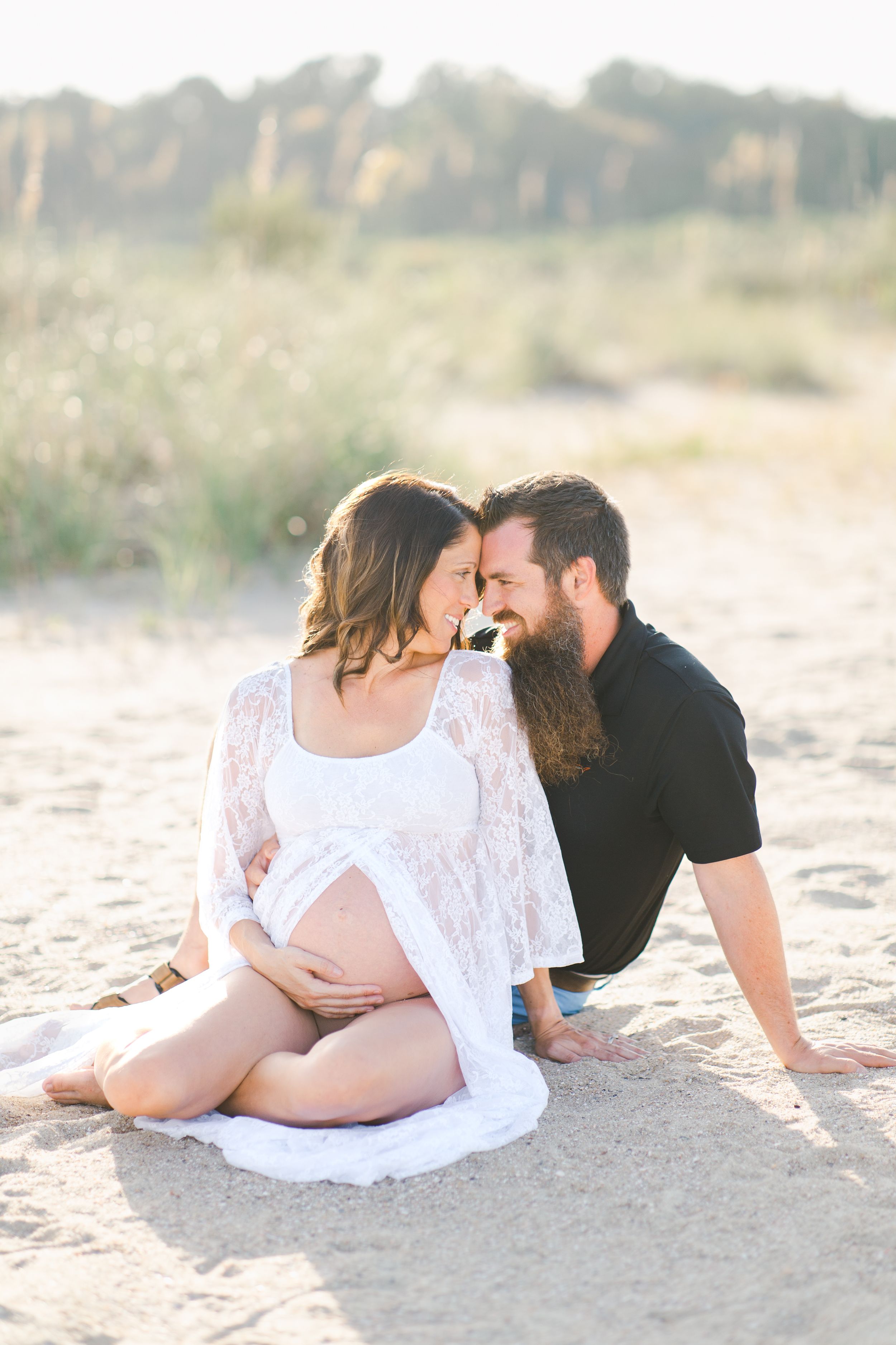 A couple in white and black outfits share intimate moments while lying on a sandy beach dunes at sunset in Hilton Head for maternity session