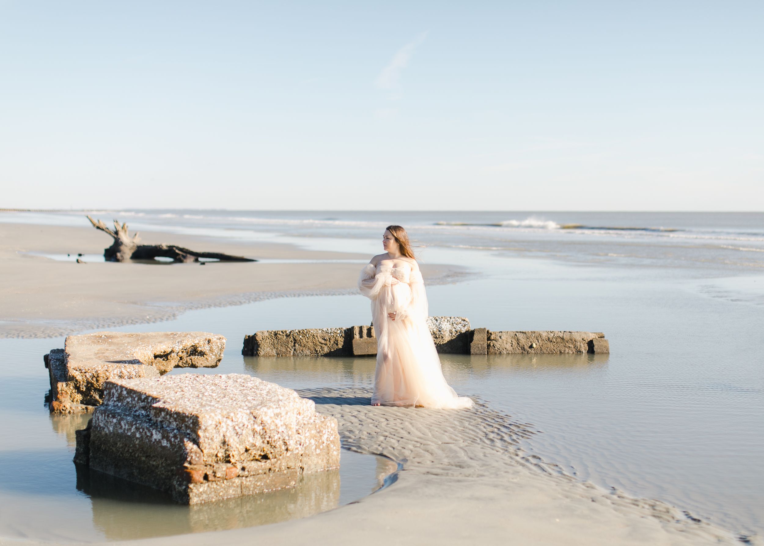 Elegant wedding dress silhouette against a serene Hunting Island backdrop with waves and rocks at sunset in dramatic maternity photography.