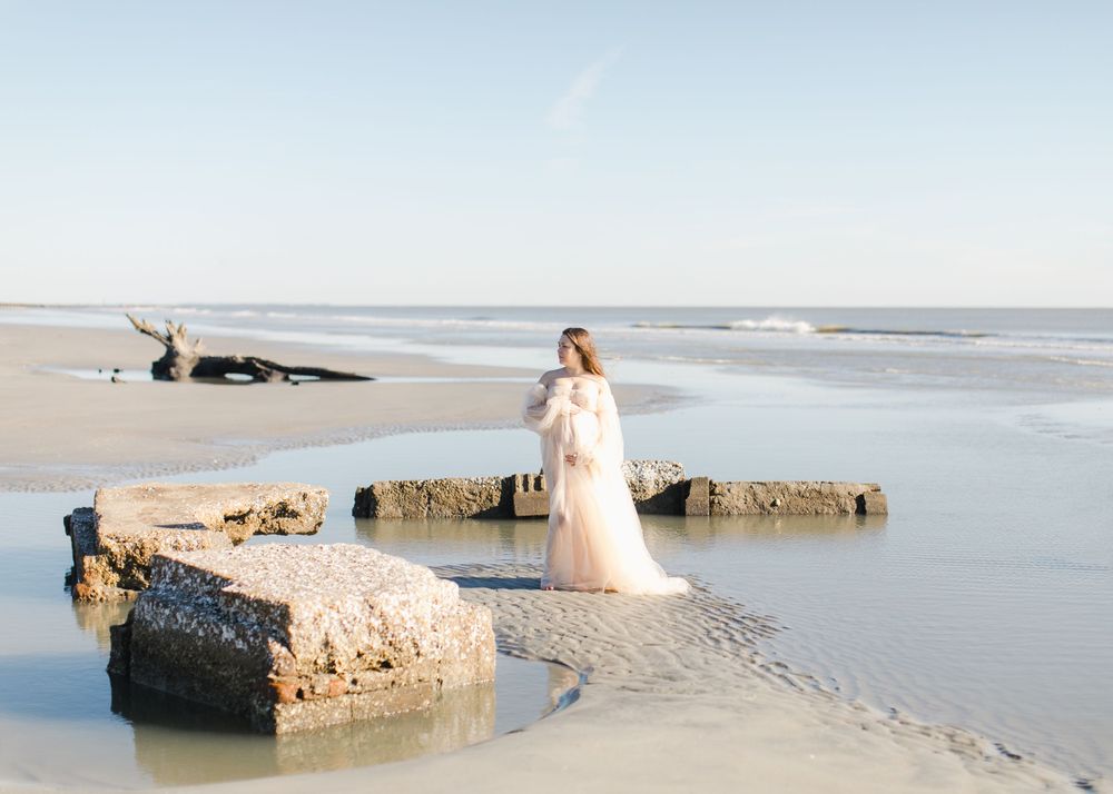 Elegant wedding dress silhouette against a serene Hunting Island backdrop with waves and rocks at sunset in dramatic maternity photography.