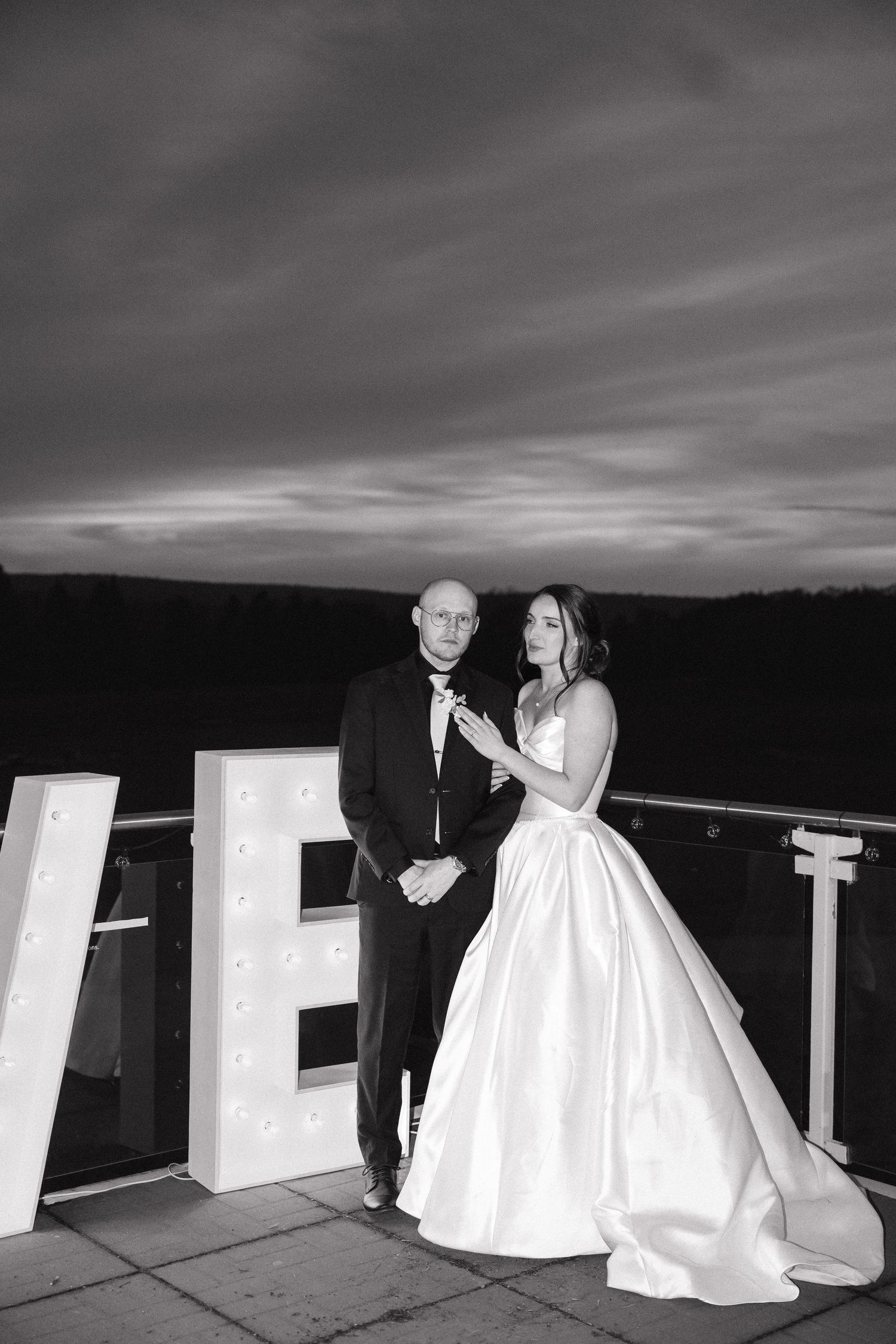 Black and white photo of bride and groom posing beside illuminated LOVE letters on an outdoor terrace at Oakfield Golf and Country Club.