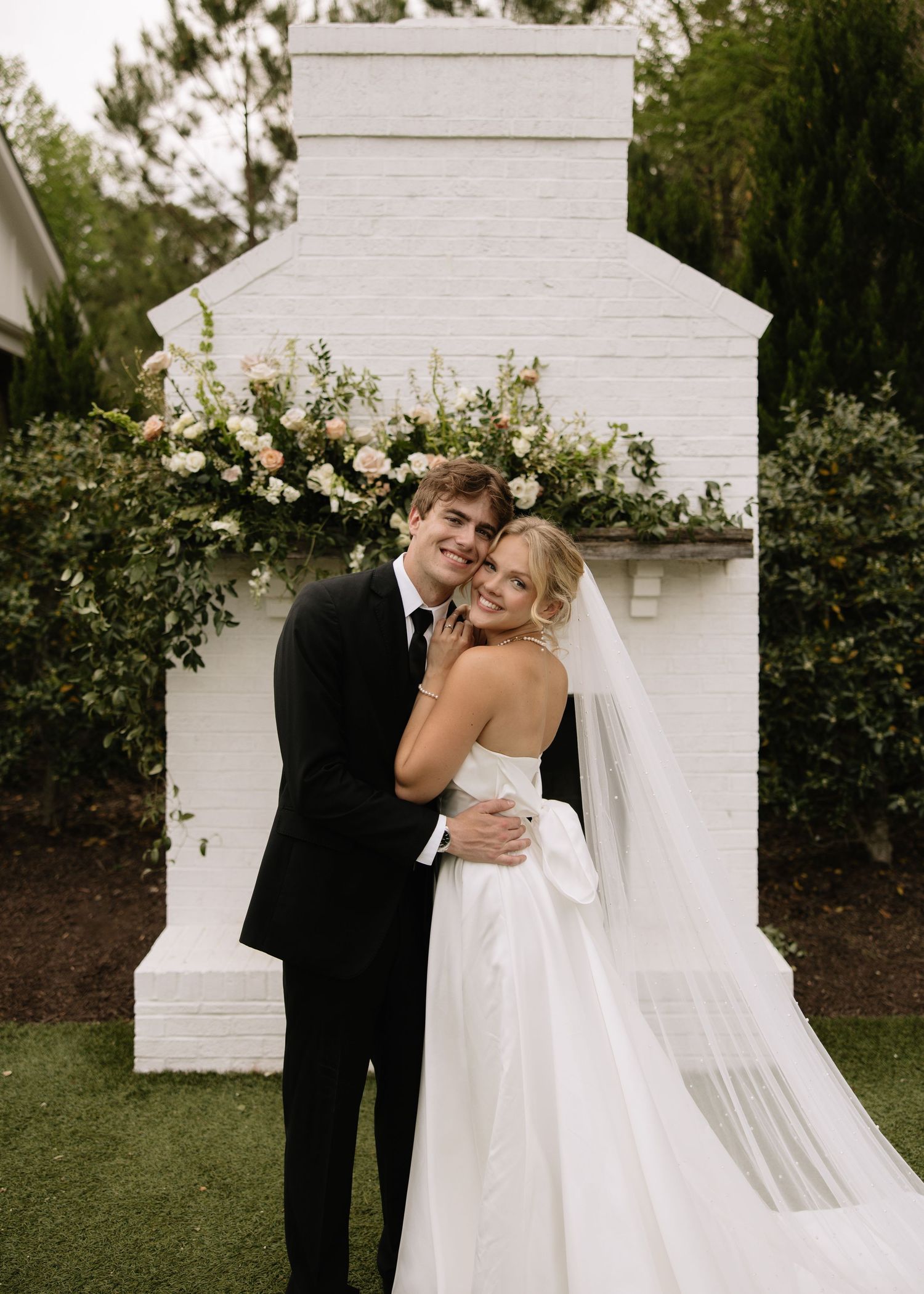 A bride and groom sharing an intimate moment against a white outdoor backdrop with flowers.