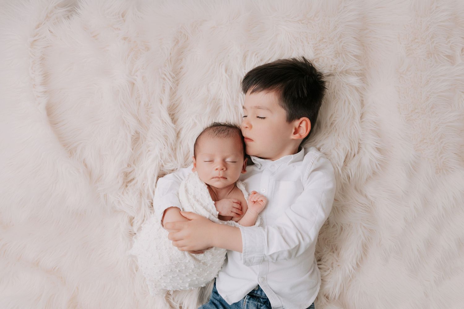 Older brother cuddling newborn baby on soft cream fur blanket. Peaceful sibling portrait captured by Fatima Photography, Basildon, Essex.
