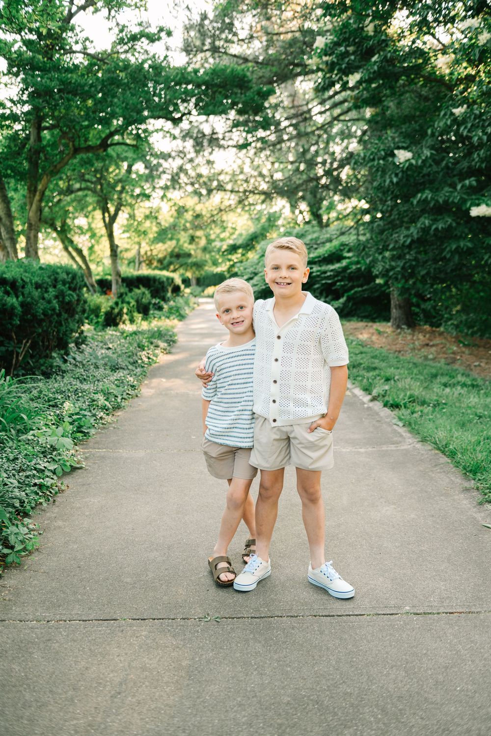 Two people walk together on a paved path surrounded by lush green trees and foliage on a sunny day.