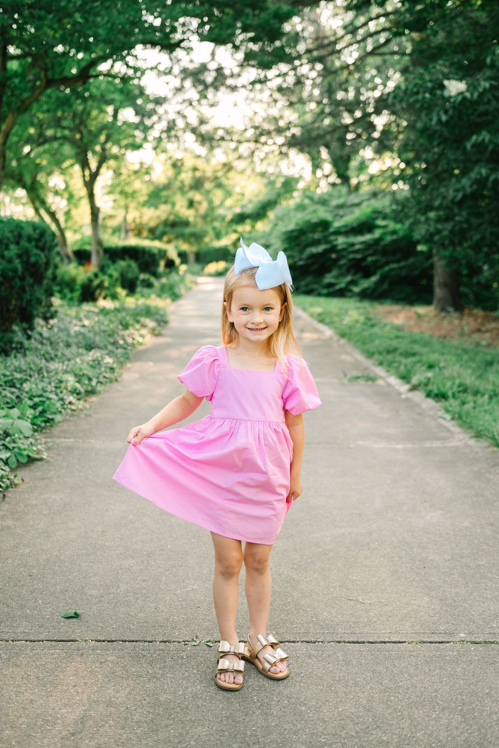 A child in a pink dress twirls playfully on a tree-lined path during a sunny day.