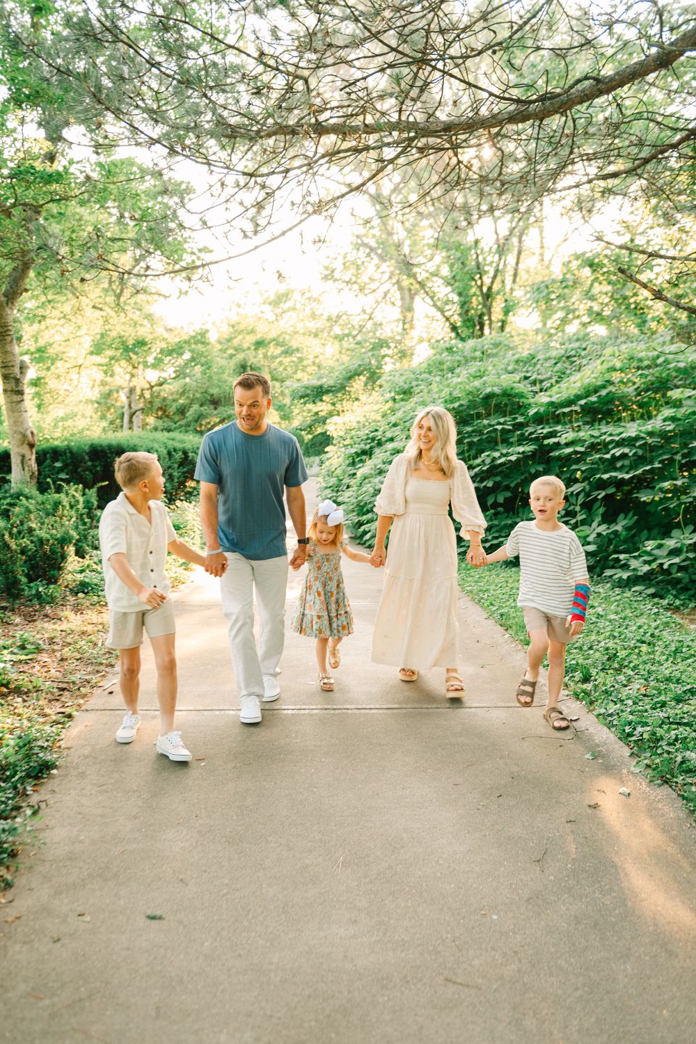 A family holds hands while walking together on a tree-lined path during a sunny afternoon.