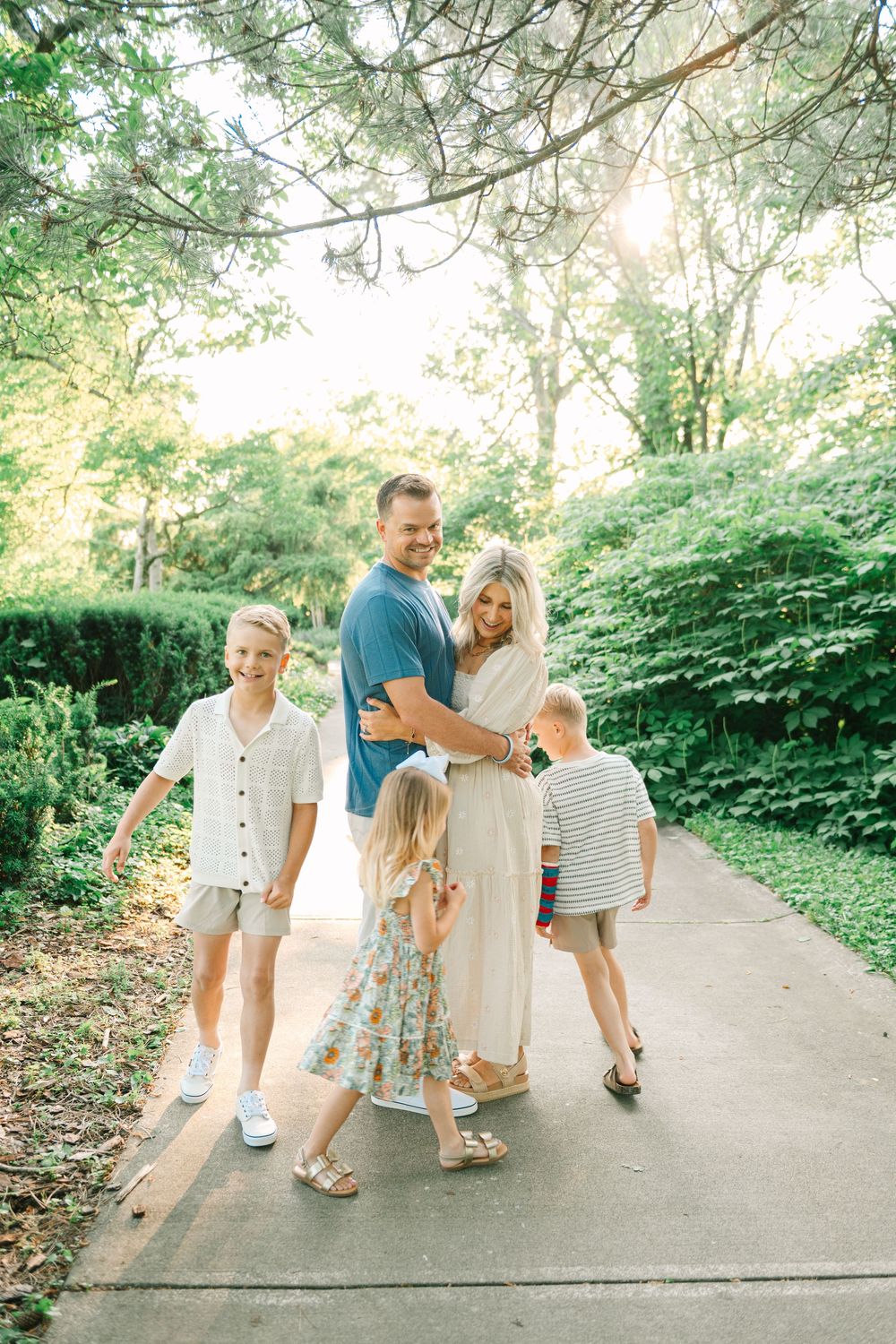 A family walks together on a sunlit garden path surrounded by lush greenery on a summer day.