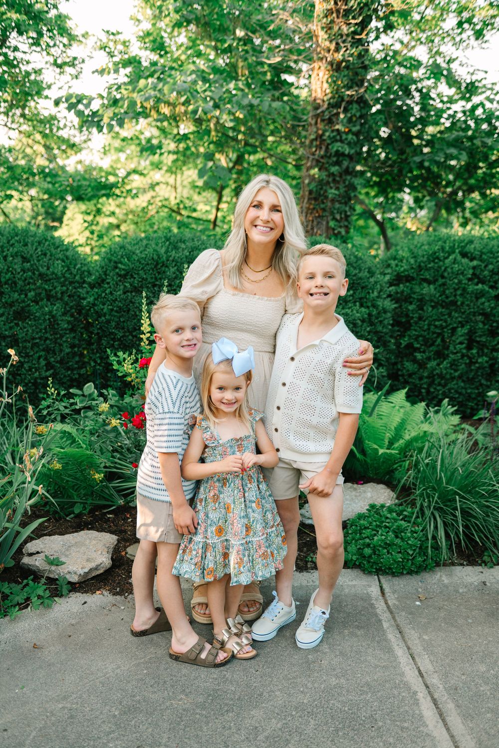 Family sits together on garden path surrounded by lush greenery and landscaping on a sunny summer day.
