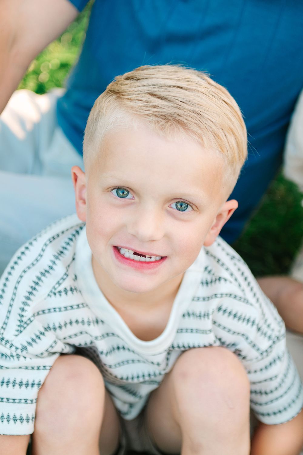 Young person in striped white shirt smiles while sitting outdoors with green foliage background.