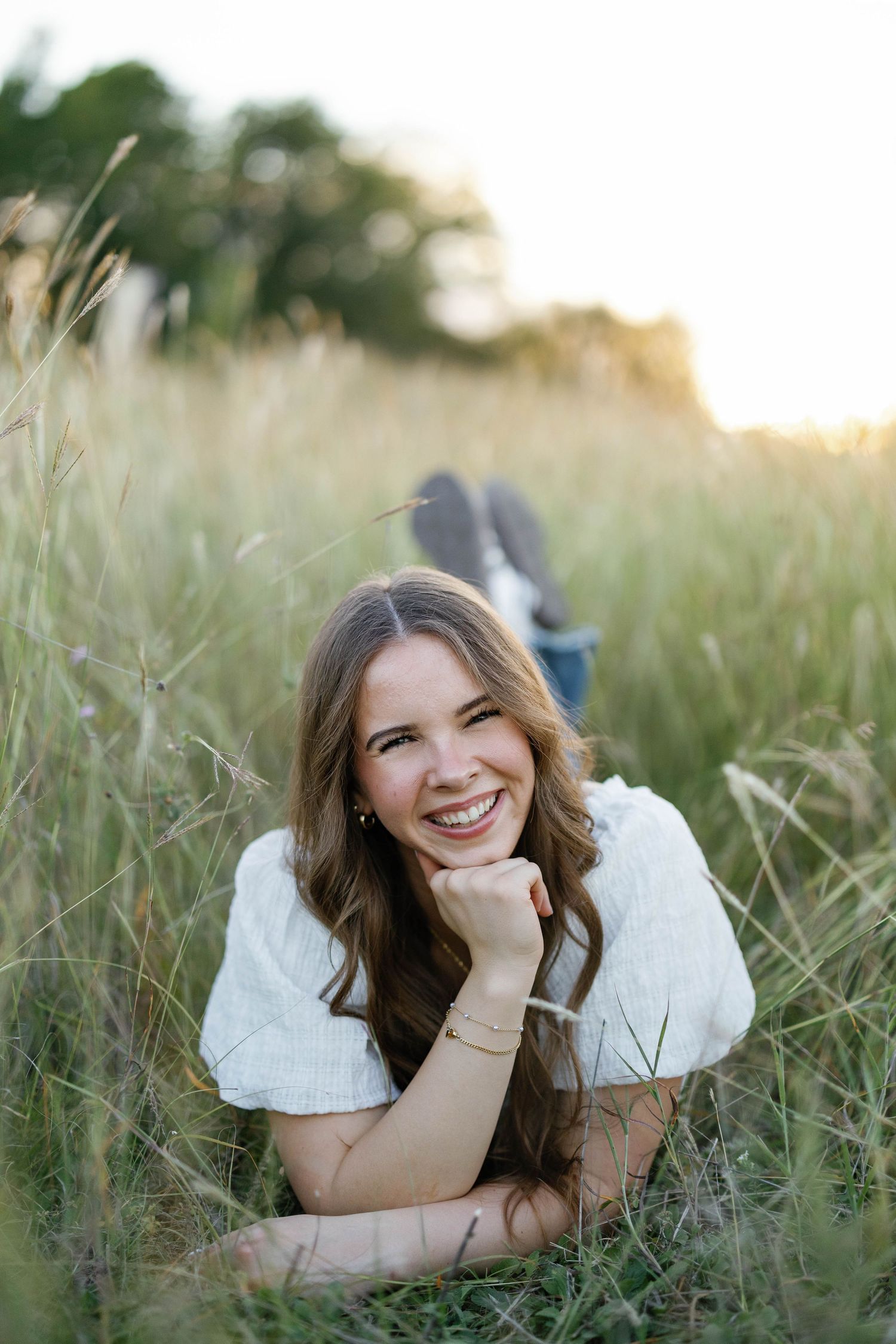 Smiling young woman in white top lying in tall golden grass field at sunset.