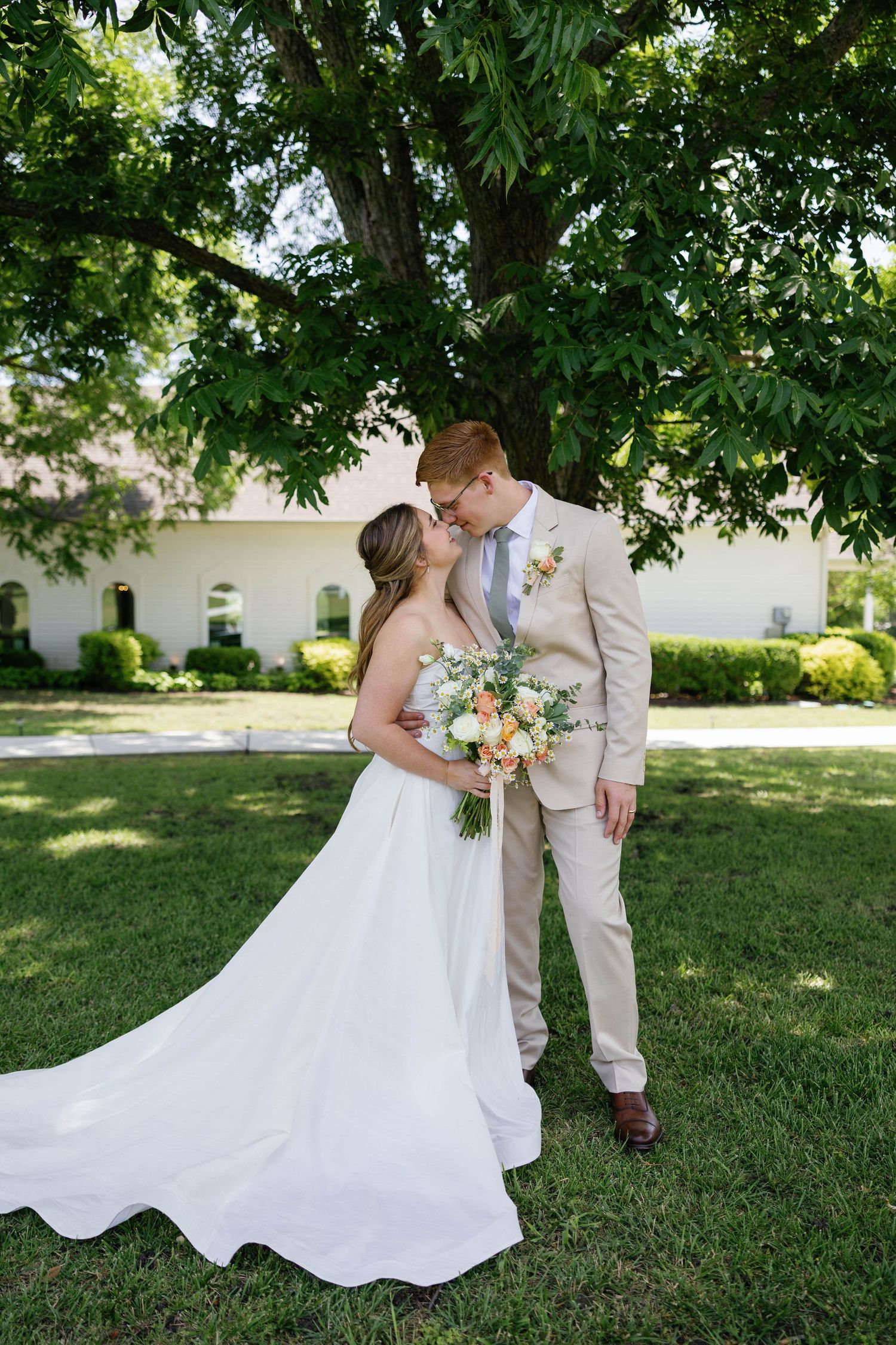 Bride and groom share a kiss beneath a large tree in a sunlit outdoor garden setting.