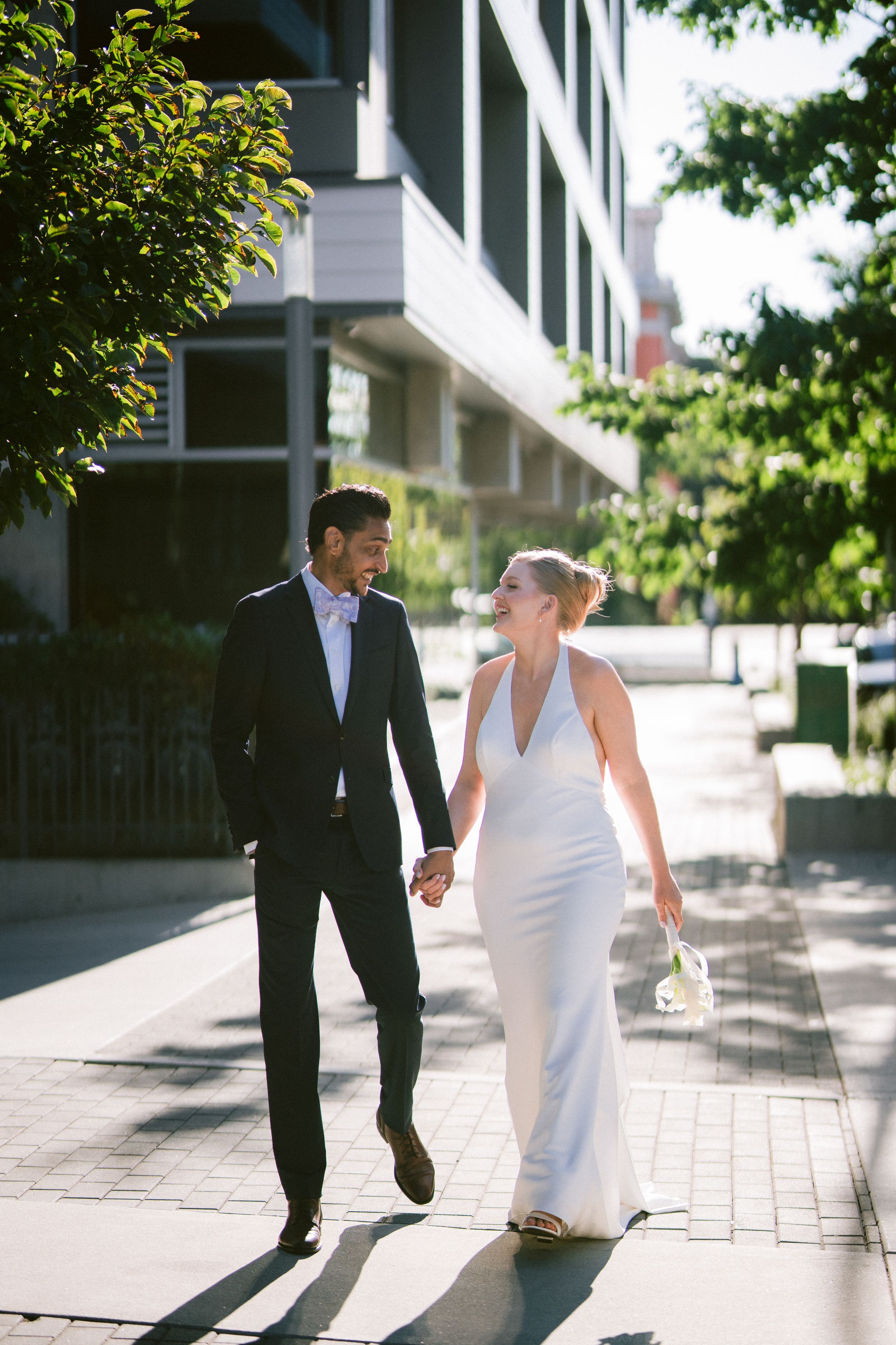 A bride and groom hold hands walking on a sunny urban sidewalk, she in a white gown, he in a black suit.