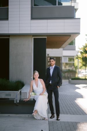 Bride in white gown sits beside groom in dark suit outside modern building for elegant wedding portrait.