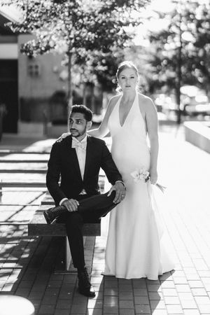 Black and white wedding photo of bride in halter gown standing behind groom seated on bench outdoors.