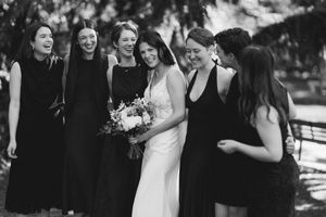 Black and white photo of a bride in white gown holding bouquet laughing with bridesmaids in black dresses outdoors.