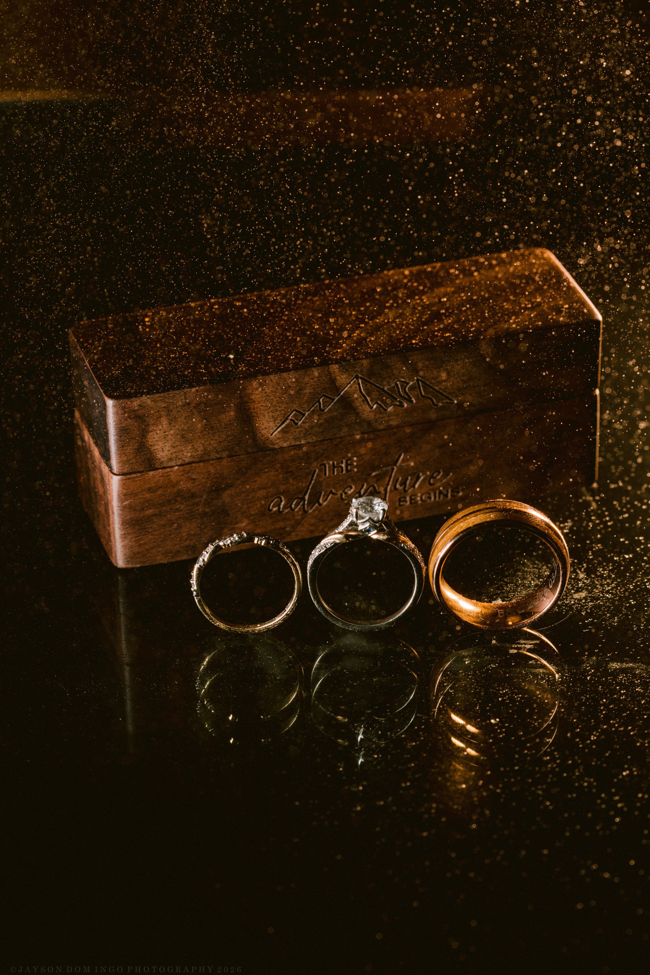 Three wedding rings displayed in front of a wooden ring box on a reflective dark surface with golden bokeh.