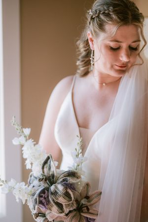 Bride in white gown holds floral bouquet with veil draped over shoulder, gazing down in soft natural light.