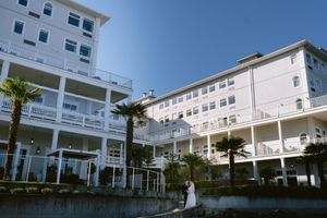 Bride and groom pose outside a grand white multi-story hotel with palm trees and balconies on a sunny day.