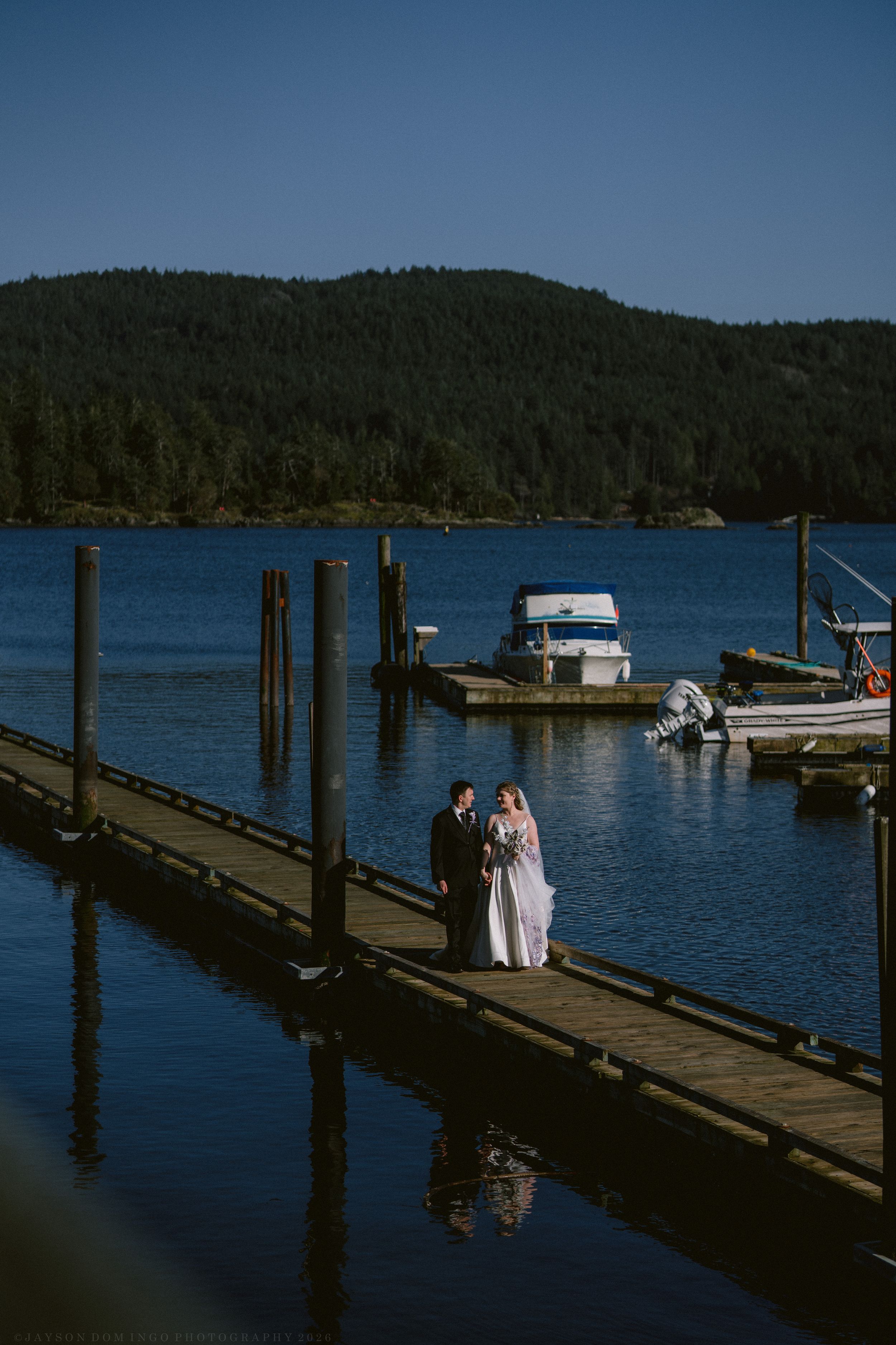 Bride and groom stand on a wooden dock overlooking a serene blue lake with boats and forested hills.