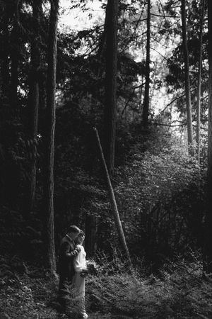 A person stands in dark moody forest reading or looking at something while surrounded by tall trees in black and white.