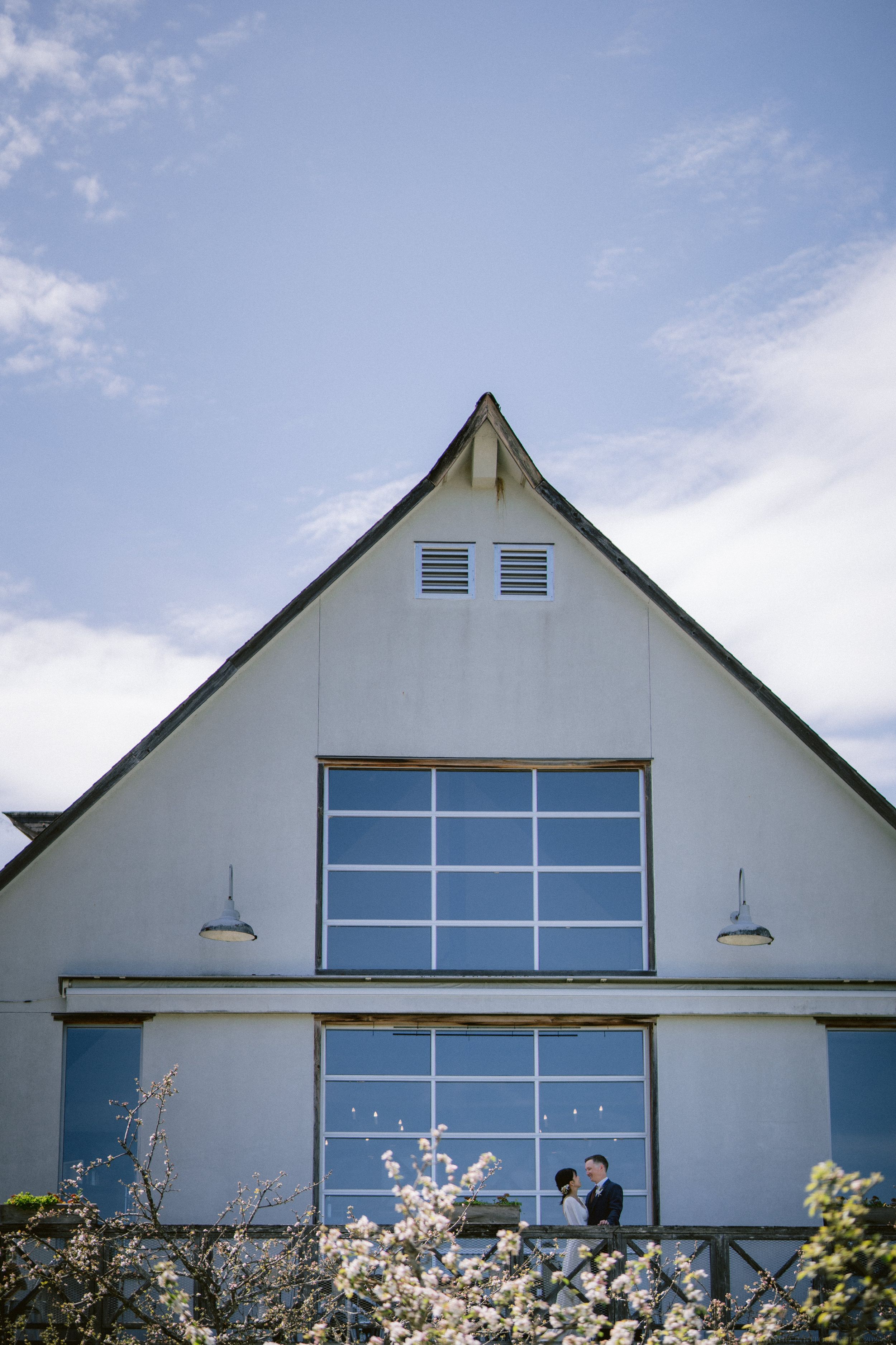 A white modern barn-style building with blue glass windows and exterior lamps, framed by blooming spring flowers.
