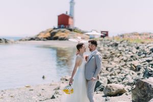 A couple poses by a lighthouse on rocky coastal shores with bright yellow flowers.