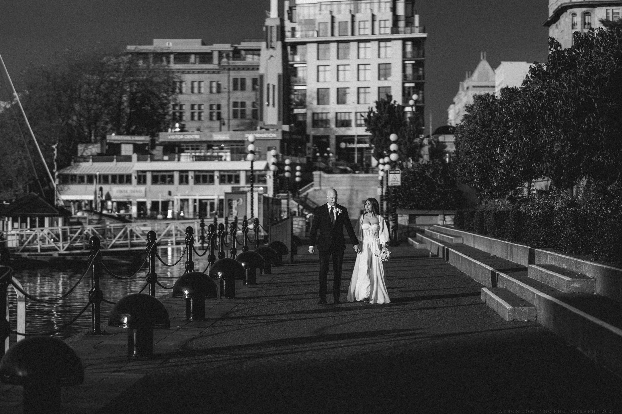 Black and white photo of a bride and groom walking on a waterfront pier with historic buildings in the background.