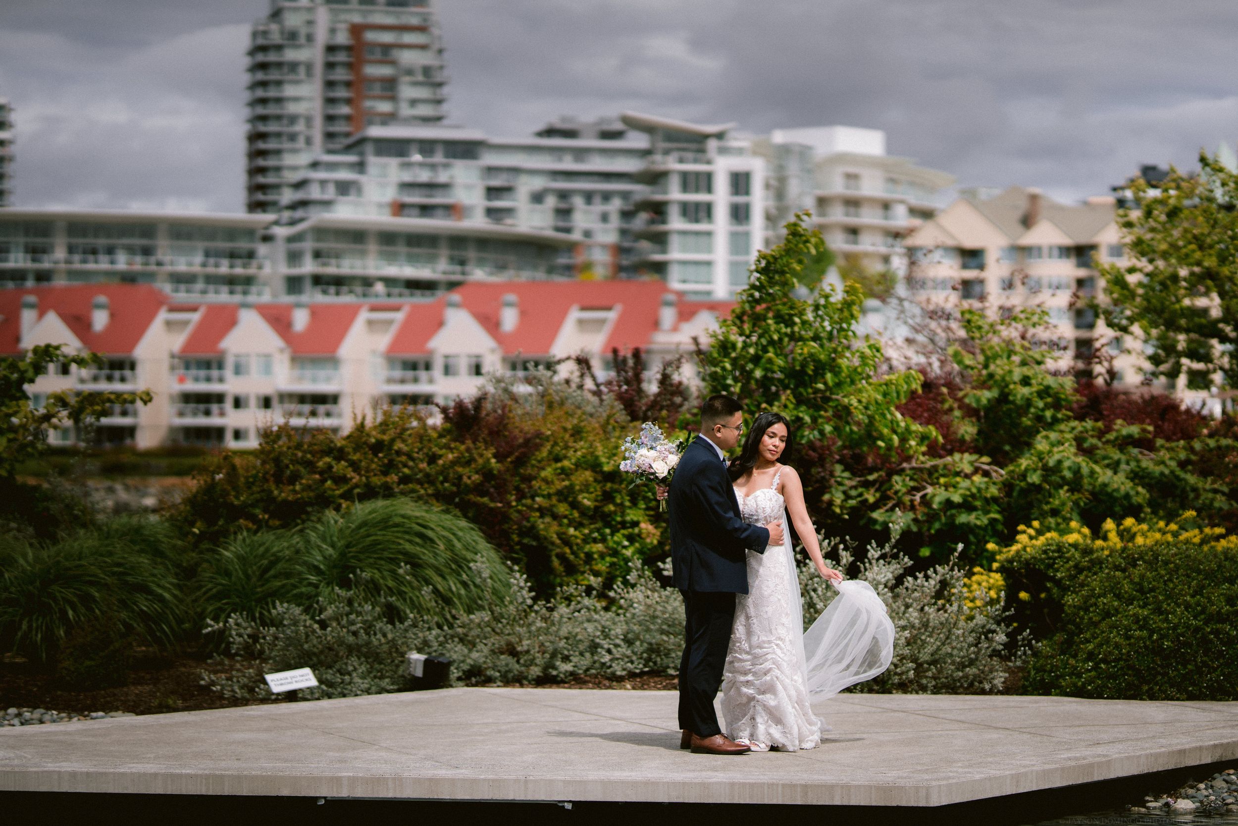 A couple in formal wedding attire embracing on a rooftop garden with city buildings in the background.