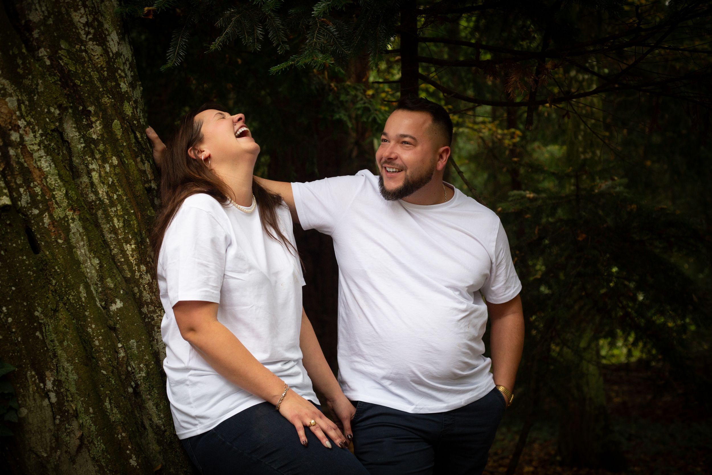 Candid moment of couple laughing together while posing by large tree trunk.