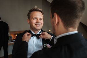 Someone helps adjust a black bowtie on a person wearing a tuxedo with boutonniere at a formal event.