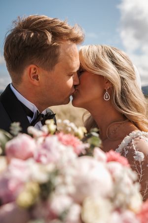 A romantic wedding moment captured with pink and white flowers in the foreground as newlyweds share a tender kiss.
