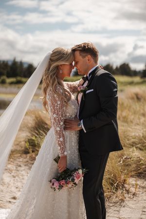 Bride and groom share intimate moment in field with flowing veil and pink bouquet on sunny day.