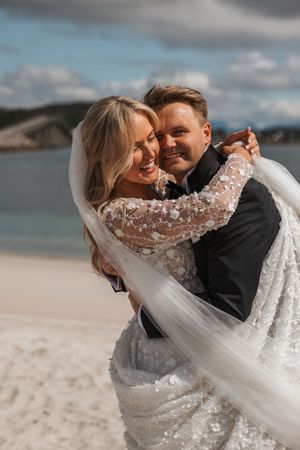 A joyful couple embraces on a scenic beach, bride wearing a lace gown with flowing veil against coastal backdrop.