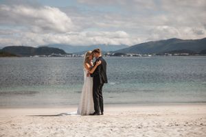 A couple in wedding attire embraces on a sandy beach with mountains and calm waters in the background.