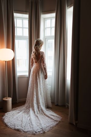 A person stands in front of bay windows wearing an intricate lace wedding gown with a long flowing train.