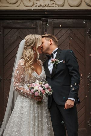 A romantic wedding couple embraces in front of dark wooden doors with a beautiful floral bouquet.