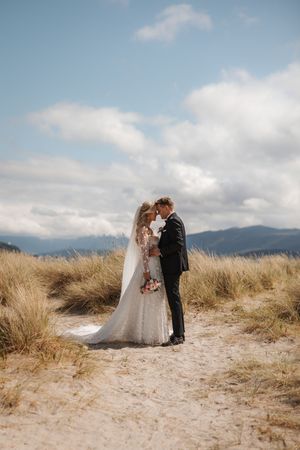 A wedding couple embraces on a hilltop with mountain views, surrounded by golden grass and dreamy clouds.