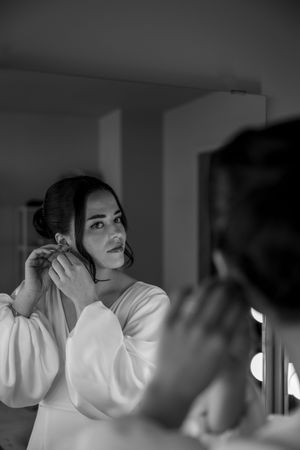 Black and white photo of someone putting on earrings while looking in a mirror, wearing a white robe or garment.