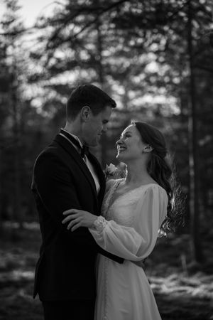 A romantic black and white wedding photo shows a couple laughing together in a forest setting.