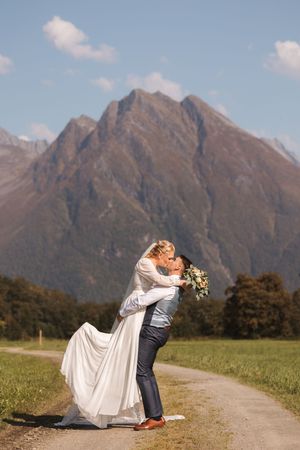 A couple in wedding attire embraces on a mountain path with majestic snow-capped peaks in the background.