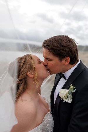 A romantic wedding kiss captured outdoors with soft veil and white boutonniere against a cloudy backdrop.
