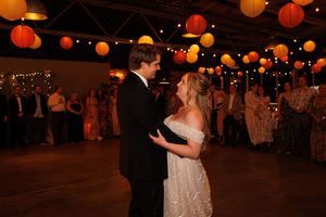 A couple dances beneath orange paper lanterns at a romantic indoor reception venue lit with warm ambient lighting.