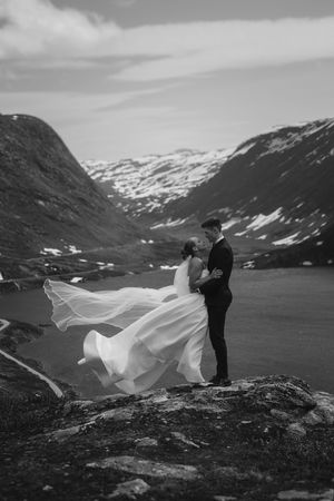 A couple stands atop a dramatic Norwegian mountain vista as a wedding dress flows elegantly in the wind.