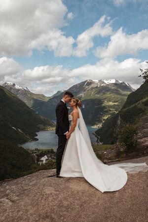 A couple embraces on a mountaintop with a stunning view of snow-capped peaks and a lake in the valley below.