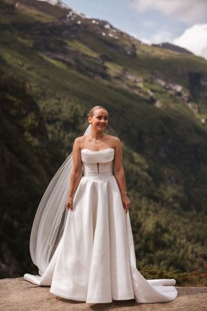 A bride in a flowing strapless white wedding dress poses on a scenic mountain overlook with a dramatic backdrop.