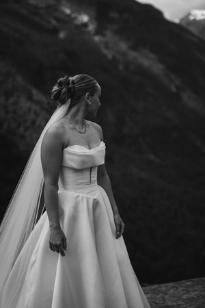 A bride in a classic white off-shoulder wedding gown with flowing veil stands against a dramatic mountain landscape.