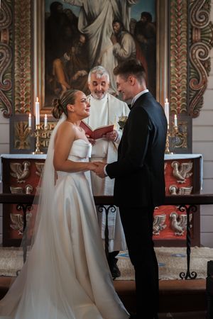 A traditional wedding ceremony takes place at the altar of a church with candlelight and religious artwork in the background.