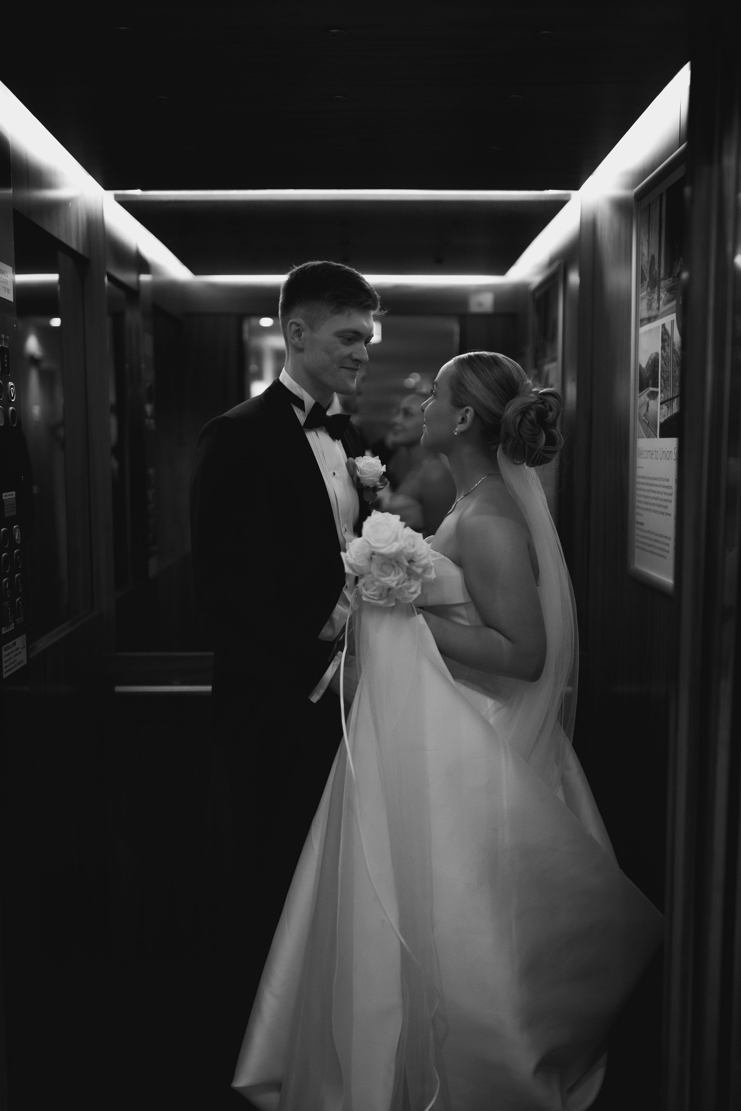 Elegant black and white wedding photograph of newlyweds sharing intimate moment in dimly lit hallway with flowers.