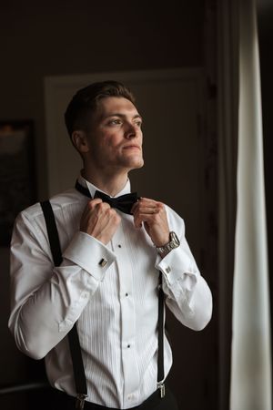 A person adjusts their bow tie while wearing a crisp white tuxedo shirt and suspenders in moody lighting by a window.
